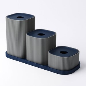 May include: A set of three gray and navy blue containers of varying heights. Each container has a ridged texture and a square top with a circular opening. The containers sit on a navy blue tray.