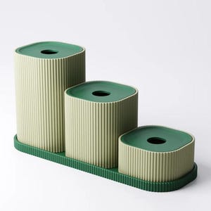 May include: A set of three green and beige containers with a ribbed texture. The containers are arranged in a stepped pattern on a green tray. Each container has a dark green, square-shaped lid with a circular opening.