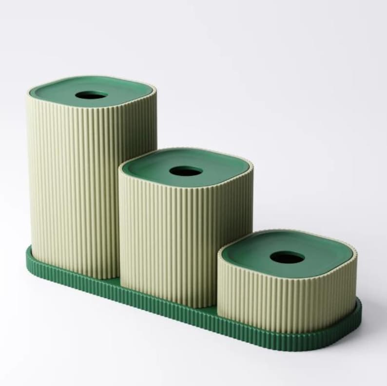 May include: A set of three green and beige containers with a ribbed texture. The containers are arranged in a stepped pattern on a green tray. Each container has a dark green, square-shaped lid with a circular opening.