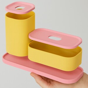 May include: A set of three-piece storage containers in yellow and pink. The containers have a ribbed texture and are rectangular and cylindrical. The lids have a rounded rectangular opening. The set is held by a hand.