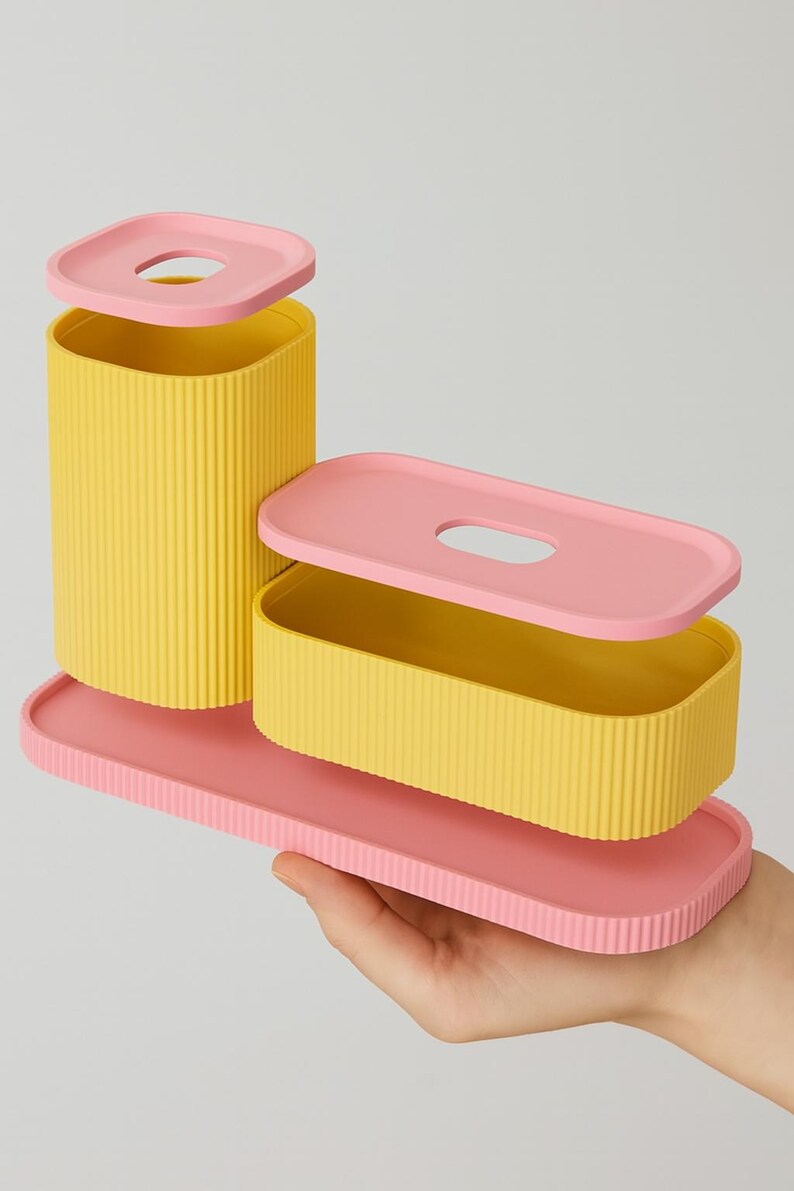 May include: A set of three-piece storage containers in yellow and pink. The containers have a ribbed texture and are rectangular and cylindrical. The lids have a rounded rectangular opening. The set is held by a hand.