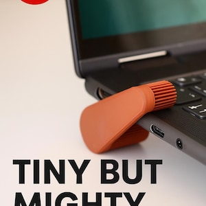 May include: An orange laptop accessory rests on a black laptop keyboard. The accessory has a curved shape with a textured, ridged end. The text "TINY BUT MIGHTY" is displayed in bold, black letters.