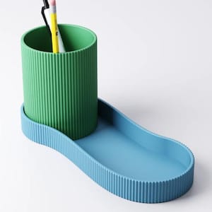 May include: A two-tone desk organizer in green and blue. The green cylindrical cup has vertical ribbed detailing and holds two pencils and a pen. The blue base has a matching ribbed edge and a curved, elongated tray.