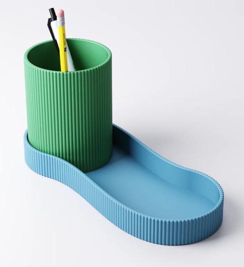 May include: A two-tone desk organizer in green and blue. The green cylindrical cup has vertical ribbed detailing and holds two pencils and a pen. The blue base has a matching ribbed edge and a curved, elongated tray.