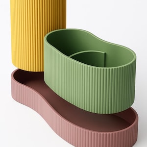 May include: A modern desk organizer set with a minimalist design. The set includes a yellow cylindrical pen holder, a green oval-shaped pen holder, and a pink storage tray. All pieces have a ribbed texture.