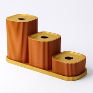 May include: A set of three orange tissue box holders with a ribbed texture, each with a gold-colored top and a circular opening. The boxes are arranged on a matching gold tray.
