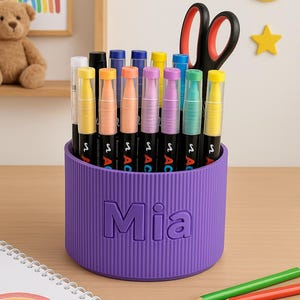 May include: A purple pen holder with the name "Mia" embossed on the front, filled with colourful markers and a pair of scissors. A notebook with a rainbow drawing and coloured pencils are on the table.