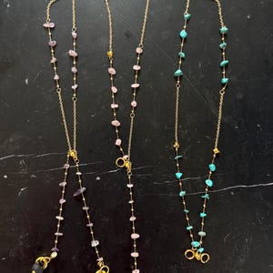 May include: Three gold chains with different colored gemstone beads. The chains have two gold rings at the bottom. The chains are on a black surface.