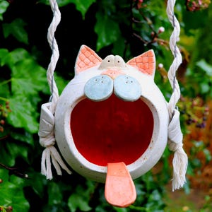 May include: A whimsical, white ceramic cat-shaped birdhouse with a wide open mouth and a red interior. The cat has blue eyes, peach-colored ears, and a tongue sticking out. It hangs from a tree branch by a white rope.