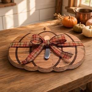 May include: A pumpkin-shaped wooden serving board with a natural wood grain finish, featuring a decorative red, green, and gold plaid ribbon tied in a bow across the center. A small silver-toned metal spreader knife is tucked under the ribbon.