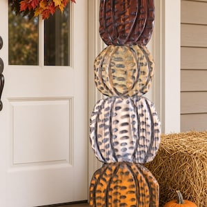May include: A vertical stack of four carved wooden pumpkins featuring a textured, distressed finish. The top pumpkin is dark brown, the second is tan, the third is light gray, and the bottom pumpkin is orange, all with dark brown accent markings.