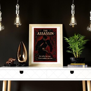 May include: A framed print titled "THE ASSASSIN" with a romantic illustration of a couple. The print is on a white table with decorative items and a plant. The text "He kills for me. Then makes me tea." is below the illustration.