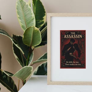 May include: Framed print featuring "The Assassin" with a romantic illustration of a couple. The artwork is set against a dark red background with floral accents. The text reads "He kills for me. Then makes me tea."