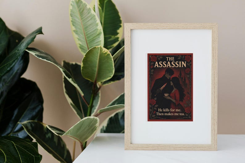 May include: Framed print featuring "The Assassin" with a romantic illustration of a couple. The artwork is set against a dark red background with floral accents. The text reads "He kills for me. Then makes me tea."