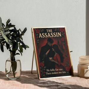 May include: A framed print with the title "The Assassin" features a romantic illustration of a couple. The artwork is set against a red background with rose accents. The text "He kills for me. Then makes me tea." is also included.