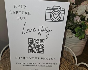 May include: A white rectangular sign with black text and a camera icon featuring a heart. The text reads HELP CAPTURE OUR Love story, SHARE YOUR PHOTOS, SCAN THE QR CODE WITH YOUR PHONE AND UPLOAD TO OUR SHARED ALBUM, Amanda & Graham 23/08/25.