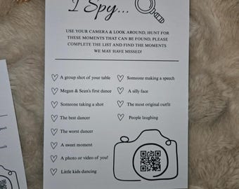 May include: A white card with the text "I Spy..." and a list of prompts for a wedding or event. The card includes a camera graphic with a QR code and a magnifying glass. The prompts include "Megan & Sean's first dance" and "A photo or video of you!"