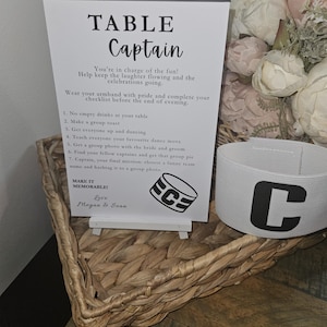 May include: A white sign with the words "TABLE Captain" and a checklist for the evening's activities. A white armband with a large black "C" is next to the sign. The sign is in a woven basket.