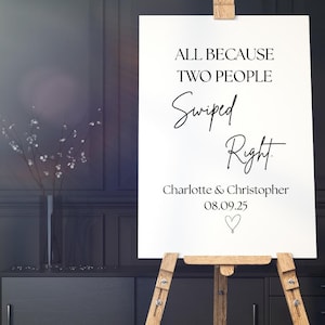 Pode incluir: Uma placa branca em um cavalete de madeira com o texto "ALL BECAUSE TWO PEOPLE Swiped Right". Abaixo do texto estão os nomes "Charlotte & Christopher" e a data "08.09.25" com um símbolo de coração. A placa está em uma sala com móveis escuros.