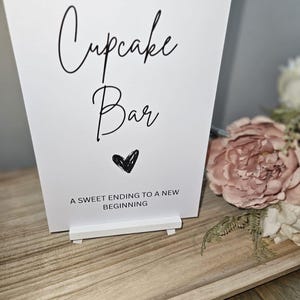 May include: A white sign reading "Cupcake Bar" in cursive script, with a black heart and the words "A sweet ending to a new beginning." The sign is on a white stand, with pink flowers in the background.