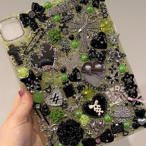 Może przedstawiać: Ozdobne etui na telefon z jasnozielonym tłem, ozdobione różnymi ozdobami. Zawiera czarne kokardy, srebrne krzyże, serca, gwiazdy i zielone akcenty. Etui ma błyszczące wykończenie i różnorodne tekstury.