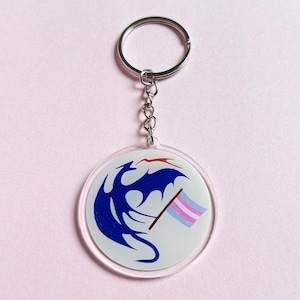 Peut inclure: Un porte-clés en acrylique transparent avec un anneau et une chaîne en métal argenté. Le porte-clés présente un motif de dragon bleu et un drapeau de la fierté transgenre sur fond blanc. Le fond est rose clair.