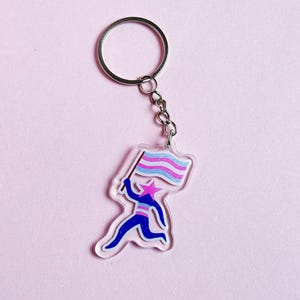 Puede incluir: Un llavero de acrílico transparente con una figura corriendo que sostiene una bandera con rayas rosas, celeste y blancas. La figura es azul con una cabeza en forma de estrella. El llavero tiene un anillo y una cadena de metal plateado.