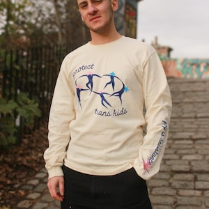 Peut inclure: T-shirt crème à manches longues avec l'inscription « protect trans kids » et un graphisme de personnages stylisés. La manche porte l'inscription « hope is resistance ». La personne porte un pantalon noir.
