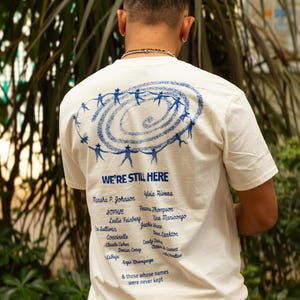May include: An off-white short-sleeve t-shirt featuring a blue graphic on the back. The design shows a spiral of figures holding hands, the text "WE'RE STILL HERE," and a list of names including Marsha P. Johnson and Sylvia Rivera.