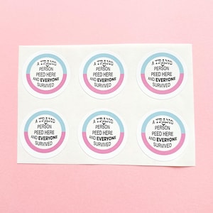 Puede incluir: Una hoja de seis pegatinas redondas. Cada pegatina tiene un diseño de rayas horizontales en azul claro y rosa. El texto de cada pegatina dice: "A TRANS PERSON PEED HERE AND EVERYONE SURVIVED."