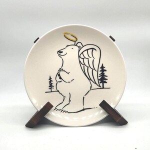 May include: A cream-colored ceramic plate featuring a whimsical illustration of a bear with angel wings and a halo. The bear is drawn in black ink, with two small pine trees on the plate. The plate is displayed on a wooden stand.