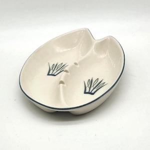 May include: Cream-coloured ceramic dish with a divided design and a dark blue trim. The dish features two stylized blue plant-like designs and two small holes. A decorative piece for serving snacks or displaying small items.