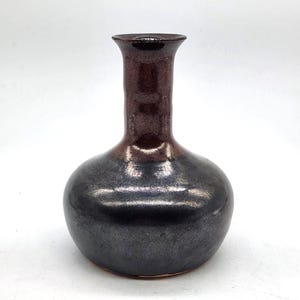 May include: A ceramic vase with a bulbous base and a narrow, flared neck. The body features a metallic, dark charcoal-grey glaze, while the neck transitions into a deep, glossy reddish-brown finish. The surface has a smooth, reflective texture.