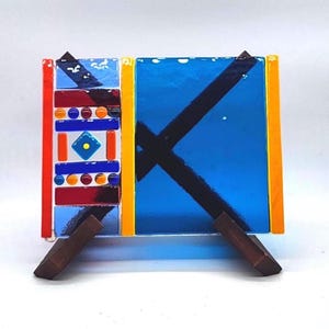 Puede incluir: Un colorido panel de vidrieras con un diseño geométrico. El panel presenta un fondo azul vibrante con una cruz negra, bordeado por amarillo y naranja. El lado izquierdo tiene un mosaico de vidrio rojo, azul y naranja. Está sostenido por un soporte de madera.