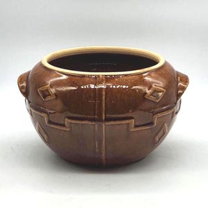 Könnte beinhalten: Eine braune Keramikschale mit glänzendem Finish. Die Schale hat eine weite Öffnung und zwei kleine Griffe auf beiden Seiten. Geometrische Muster sind auf der Außenseite geprägt. Der Rand ist hellbeige.
