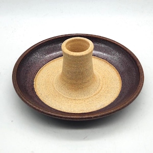 May include: A handmade ceramic incense holder with a circular base and a central cylindrical tower. The base is a deep burgundy color, while the center is a light tan. The incense holder is designed to hold incense sticks.