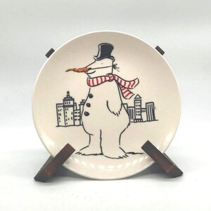 May include: A decorative ceramic plate featuring a snowman with a carrot nose, top hat, and striped scarf. The snowman stands in front of a cityscape. The plate is displayed on a wooden stand.