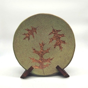 May include: A round, green ceramic plate with three brown leaf designs. The plate is displayed on a wooden stand. The plate has a textured surface and a rustic aesthetic, suitable for home decor.