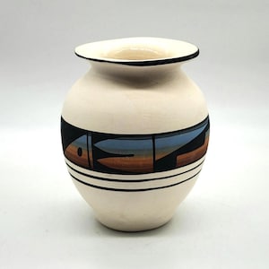 Może przedstawiać: Kremowy ceramiczny wazon o zaokrąglonym korpusie i szerokim otworze. Wazon ma czarną obwódkę i pas geometrycznych wzorów w kolorach czarnym, brązowym i niebieskim. Dwie czarne poziome linie otaczają wazon poniżej.