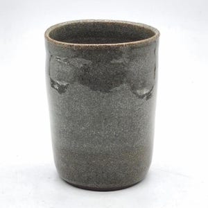 Könnte beinhalten: Ein handgefertigter, zylindrischer Keramikbecher mit einer strukturierten, grauen Außenseite und einem dunkleren Rand. Der Becher hat eine glatte, glänzende Oberfläche und ein schlichtes, minimalistisches Design, geeignet für Getränke.