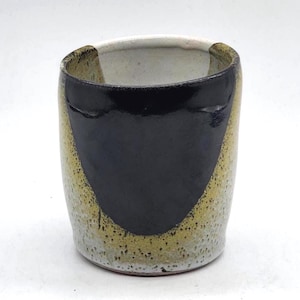 May include: A handmade ceramic tumbler with a unique design. The cup features a black, curved shape against a speckled, tan background. The rim is white, and the interior is a light beige color. This artful piece is perfect for beverages.
