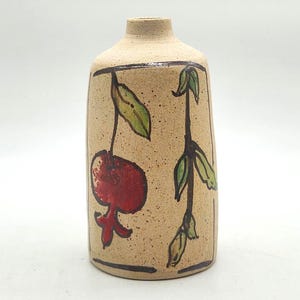 Può includere: Un vaso cilindrico in ceramica con finitura beige maculata, caratterizzato da un'illustrazione dipinta a mano di un melograno rosso e foglie verdi su un gambo, incorniciata da sottili linee nere. Il vaso ha un collo stretto e base piatta.