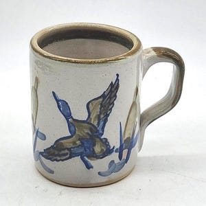 Puede incluir: Una taza de cerámica con un diseño pintado a mano de un pato volando en tonos azules y marrones. La taza tiene un fondo beige, un borde marrón y un asa curvada. La taza mide aproximadamente 10 cm de alto.