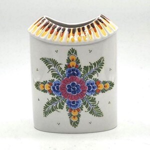 May include: A white ceramic vase with a floral design. The vase has a rectangular shape with a curved top. The floral design features blue, red, yellow, and green colors. The top rim has a brown and yellow pattern.