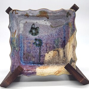 May include: A square ceramic dish with a scalloped edge, resting on a wooden stand. The dish features a purple and blue mottled glaze with abstract floral designs. The wooden stand is made of dark brown wood.