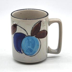 Könnte beinhalten: Ein Keramikbecher mit gesprenkelter cremeweißer Glasur und dunklem Rand. Die Außenseite zeigt ein handgemaltes Motiv aus zwei runden Früchten in Blau- und Dunkellila-Tönen, ergänzt durch braune Blätter. Er besitzt einen stabilen, eckigen Henkel.