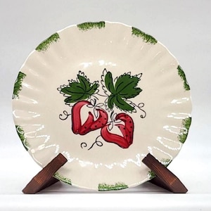 May include: Cream-colored ceramic plate with a ruffled edge, featuring a hand-painted design of two red strawberries and green leaves. The rim has a green, scalloped border. The plate is displayed on a wooden stand.