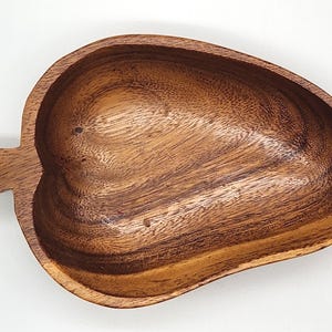 Puede incluir: Un cuenco de madera para servir con forma de hoja y asa. El cuenco está hecho de madera marrón oscuro con vetas visibles. El asa es una pequeña extensión rectangular del cuenco.