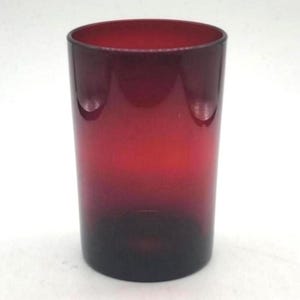 Puede incluir: Un vaso cilíndrico de color rubí. El vaso tiene un efecto degradado, que pasa de un rojo oscuro y profundo en la base a un rojo más claro y brillante en el borde. El vaso está vacío y se encuentra sobre una superficie blanca.