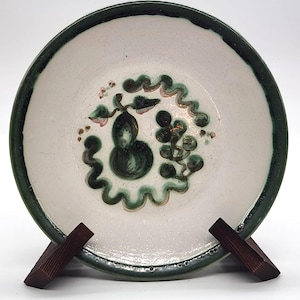 Puede incluir: Un plato de cerámica hecho a mano con interior blanco y borde verde oscuro. El plato presenta un diseño floral verde oscuro y rosa en el centro. El plato se exhibe sobre un soporte de madera.